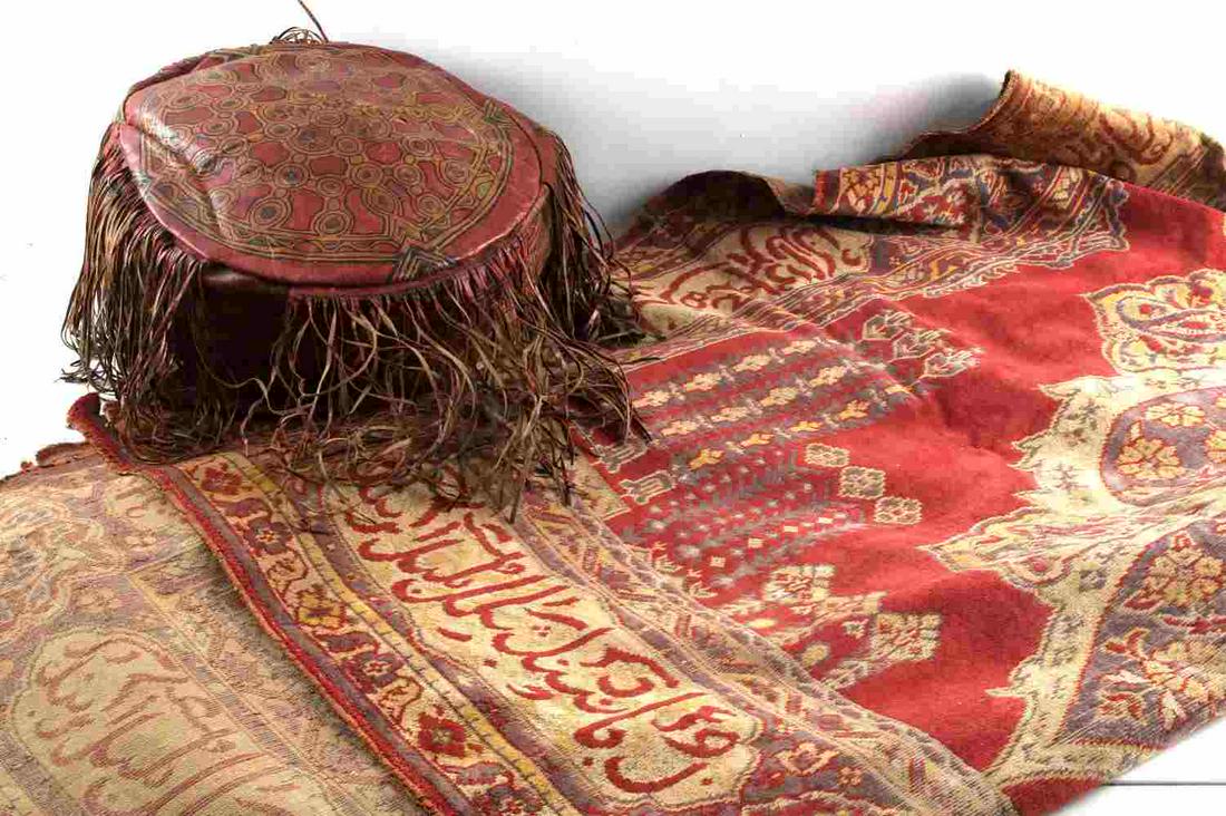 MOROCCAN FRINGED LEATHER OTTOMAN & WOOL RUG: One small-sized Moroccan fringed leather ottoman measuring 12 inches in diameter and 7 inches high. Typical to colors from Islamic cultures, the ottoman is a base red dyed leather with a concentric an