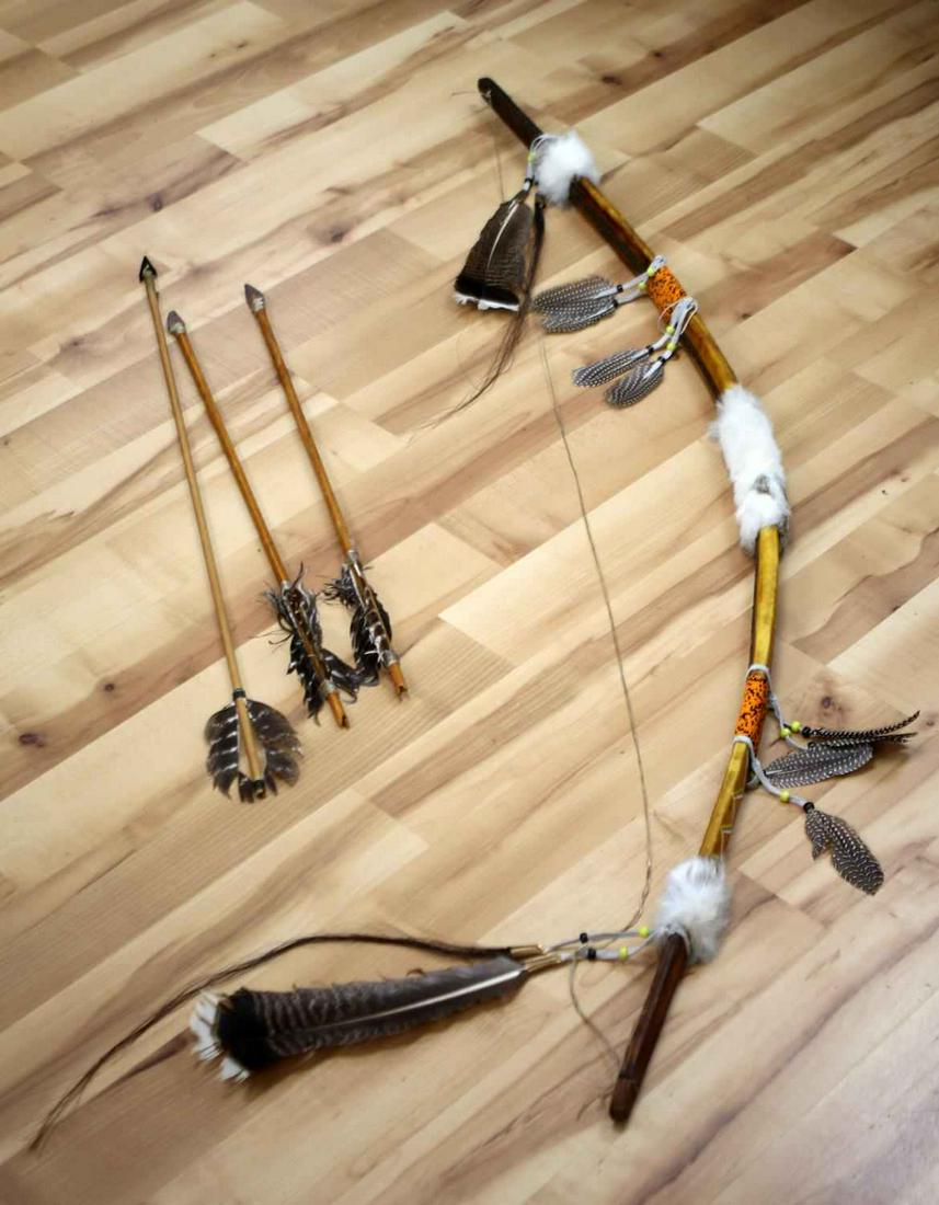 APACHE LEATHER WRAPPED ORNAMENTAL BOW & ARROWS: Wooden bow wrapped in animal hide with beaded sections, rabbit fur sections and feathers adorning, measures 60 inches long (along the curve.) Comes with three wooden arrows with arrowhead points. Very