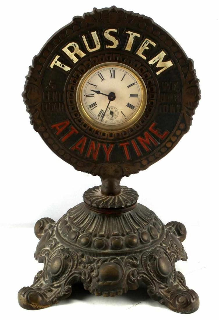 CAST IRON CIGAR CUTTER CLOCK AND ORNAMENTAL STAND: Cast iron cigar cutter clock. Clock is over-wound. ALSO MISSING THE CIGAR CUTTER. Manufactured by Waterbury Clock Co. USA. The round plaque reads "Trust Em At Any Time" and "5 Cent Cigar" of W.E. Bake