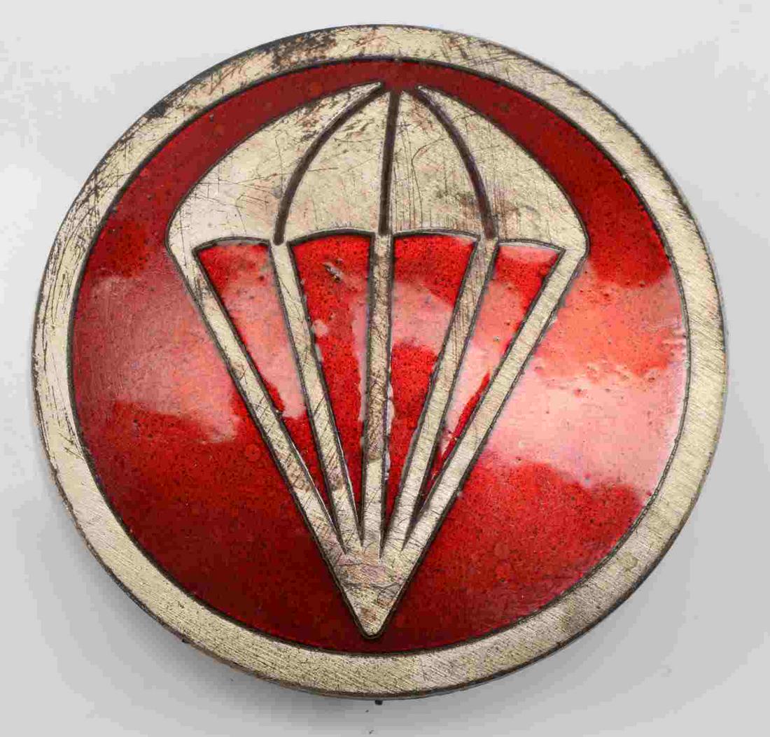 US WWII AIRBORNE PARATROOPER EM OVERSEAS CAP BADGE: Measures 1 3/4" in diameter. The front shows a parachute with a red back ground signifying artillery. Has a thin vertical English style pin back with a ‘c’ catch. Nickel construction with red enam