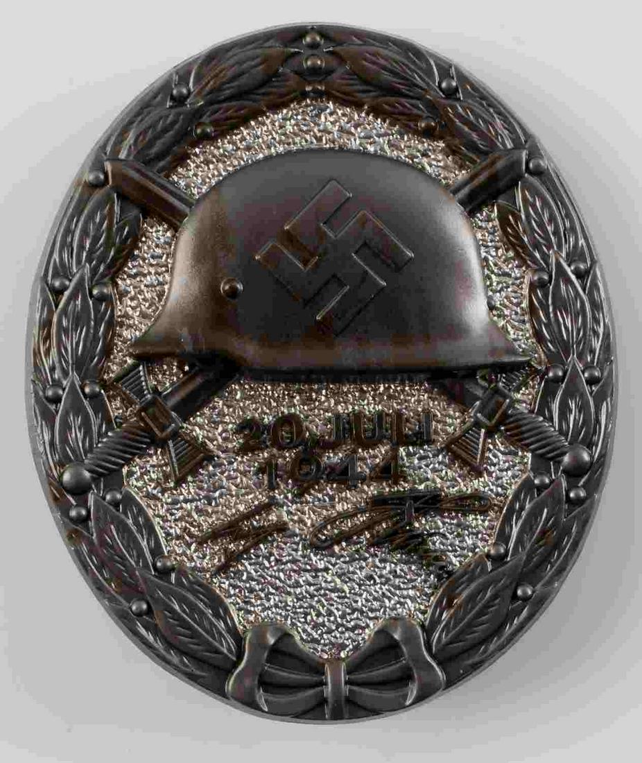 GERMAN WWII BLACK ADOLF HITLER WOUND BADGE: Measures 1 3/8” wide 1 11/16” tall. The front shows a German helmet in the center with a pair of crossed swords in the back ground. Below reads ’20 Juli 1944 Adolf Hitler’. Has an oval oak lea