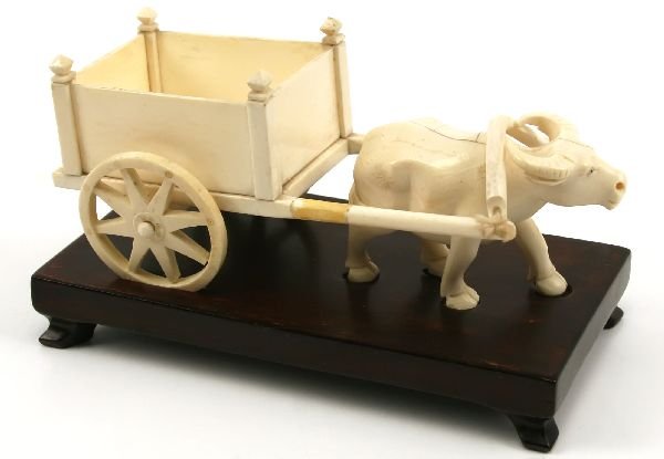 IVORY OX & WAGON FIGURINE ON WOODEN BASE: Ivory ox and wagon altogether measuring 7.25 inches x 3 inches. The wagon rests on the ox, but is not attached. Ox has minor surface cracks, left hind leg has been reattached. Both pieces rest (not at