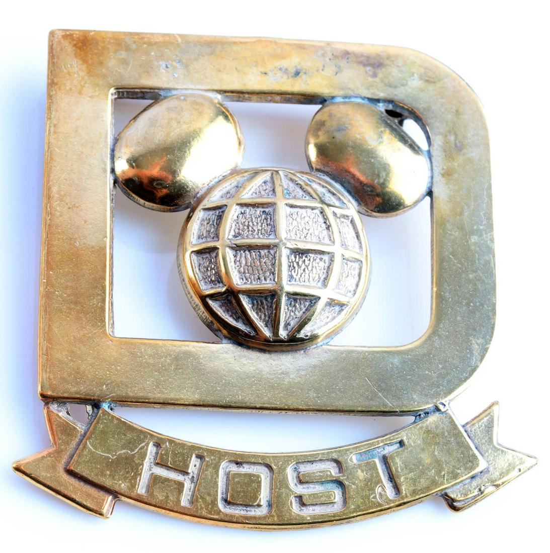 VINTAGE WALT DISNEY WORLD CAST MEMBER HOST BADGE: Vintage Walt Disney World cast member host badge. Measures 1 1/2 X 1 3/4 inches. The front shows a Mickey Mouse head in the center of a D. The ribbon below reads HOST. Has a thin horizontal pin back w
