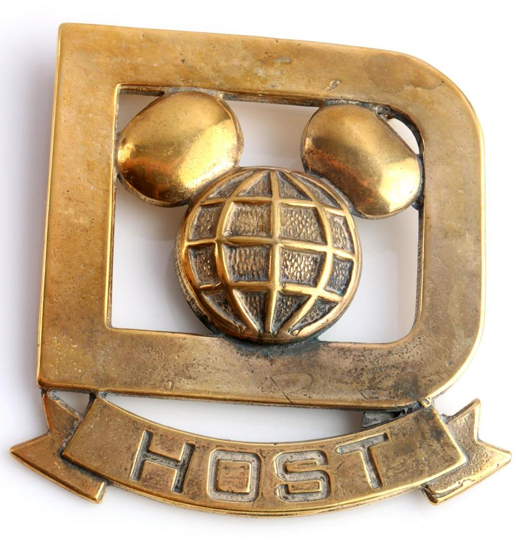 VINTAGE WALT DISNEY WORLD CASST MEMBER HOST BADGE: Vintage Walt Disney World Cast Member HOST Badge. Measures 1 1/2 X 1 3/4 inches. The front shows a Mickey Mouse likeness in the center of a D. The ribbon below reads HOST. Has a thin horizontal pin ba