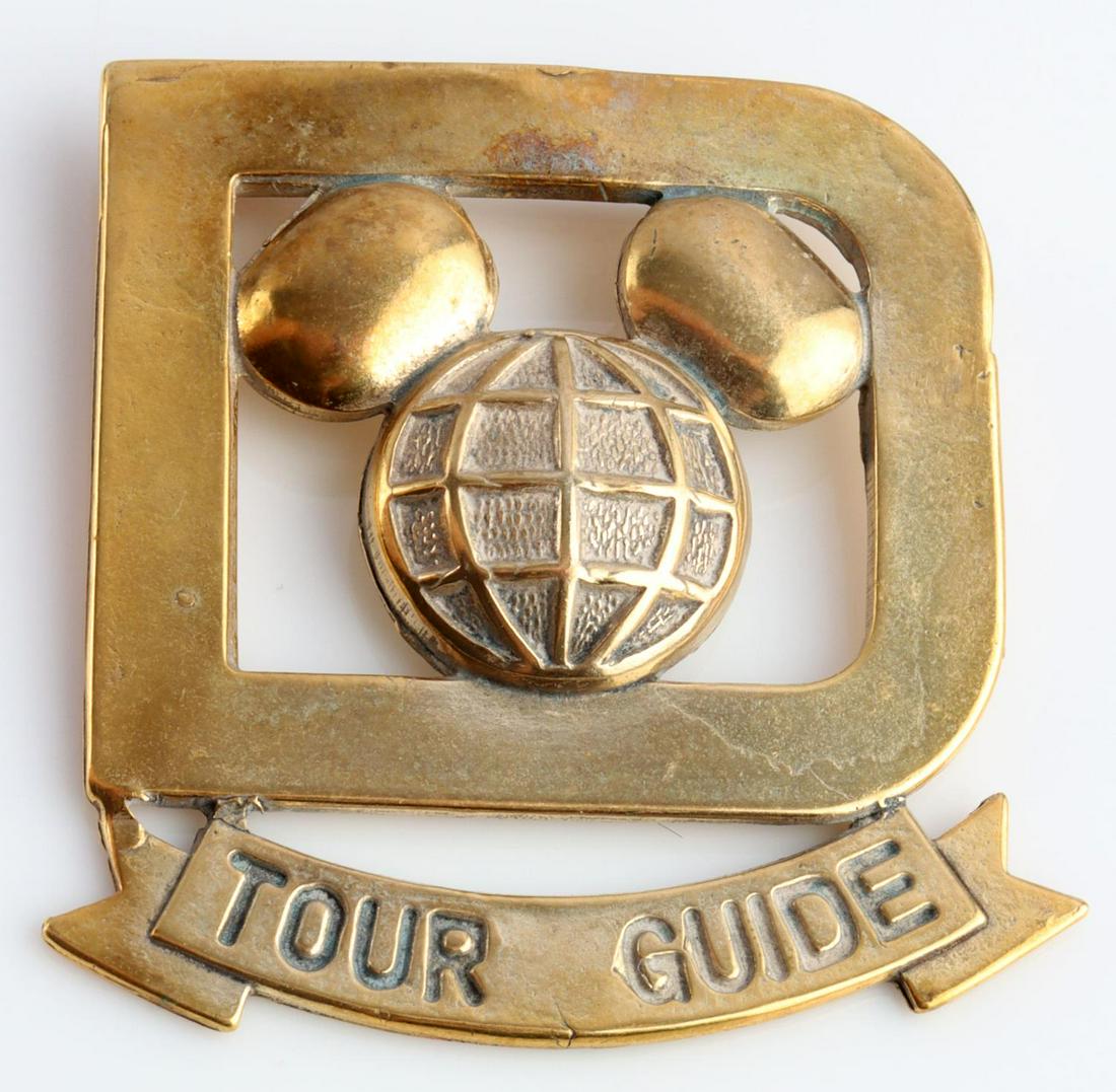 VINTAGE WALT DISNEY WORLD CAST MEMBER TOUR GUIDE: Vintage Walt Disney World Cast Member TOUR GUIDE Badge. Measures 1 1/2 X 1 3/4 inches. The front shows a Mickey Mouse likeness in the center of a D. The ribbon below reads TOUR GUIDE. Has a thin horiz