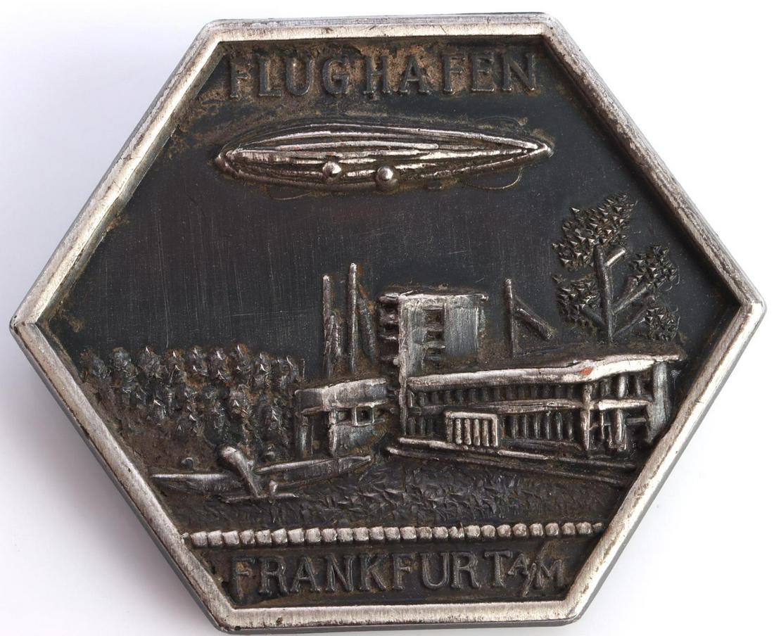 WWII GERMAN FRANKFURT ZEPPELIN AIR SHIP BADGE: German World War II Zeppelin Frankfurt Air Ship Badge. Measures 1 1/2 X 1 3/16 inches. Front reads ‘Flughafen Frankfurt A/M’ shows a Zeppelin flying over the city. Thin horizontal safety pin back.