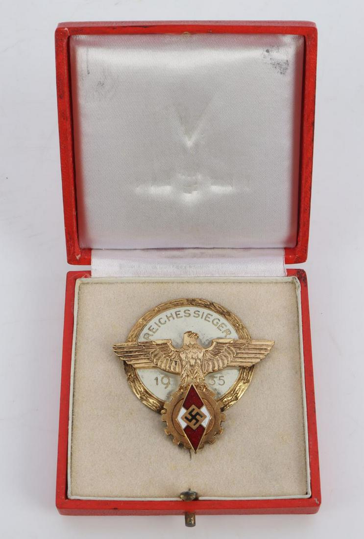 WWII GERMAN HITLER YOUTH 1935 REICHESSIEGER BADGE: Measures 1 15/16 inches wide by 2 inches tall. The front shows a German eagle clutching a swastika in his talons in the center. There is a HJ diamond surrounded by a round cog wheel at the base. All t