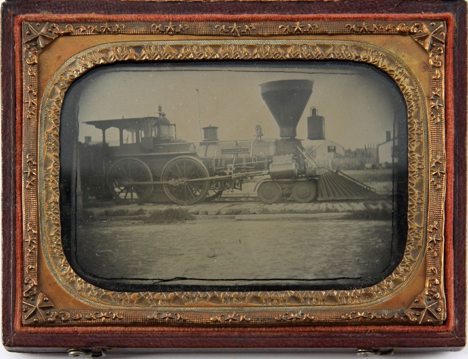 AMBROTYPE OF STEAM LOCOMOTIVE TOM GREY UNION FRAME: Ambrotype from the Civil War period in Union style frame of locomotive Tom Grey. Photograph depicts locomotive on tracks in what appears to be a train yard. The train features a name plaque which read