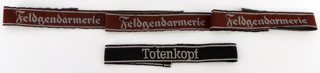 2 GERMAN CUFF TITLE SS TOTENKOPF FELDGENDARMERIE: Ex dealer stock with over $600 retail stickers still attached, the Feldgendarmerie is unissued with multiples, each very good original, non-fluorescing condition.