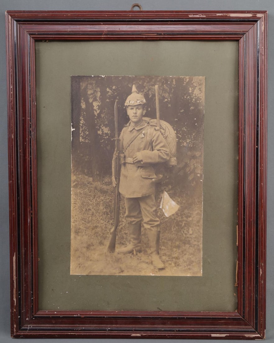 PORTRAIT OF WWI SOLDIER WITH FLOWERS IN FOREST: Period framed print photograph in wooden frame (no glass), measuring 23 X 18 inches, depicting soldier with Mauser rifle at his side standing in a forest. Soldier has flowers on helmet and lapel that 