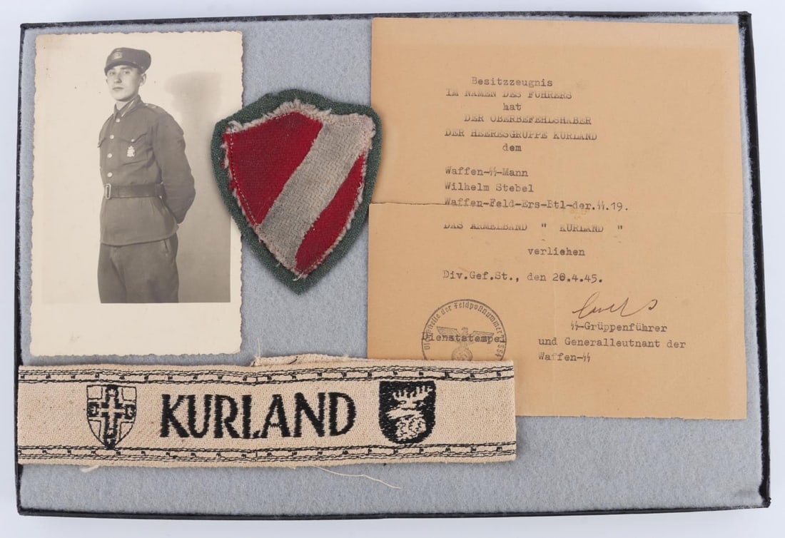 WWII LATVIAN SS KURLAND CUFFTITLE AND PATCH: Comes with document for Kurland cuff title issue. Both patch and cuff title are non fluorescing. Riker mount measures 8 1/4 X 12 1/4 inches. Very good condition. This lot has a reserve.