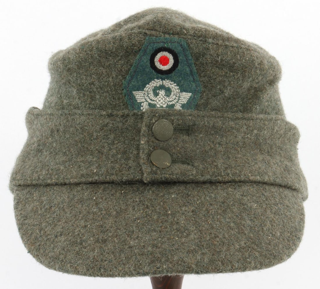 WWII GERMAN THIRD REICH POLICE M43 WOOL FIELD CAP: Police field cap in feldgrau wool. Ear flaps affixed in front with painted pebbled steel buttons. Embroidered rosette above police eagle patch machine-sewn to front. Interior has grey twill liner. No 