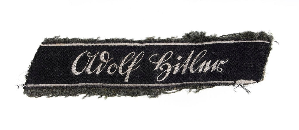 LSSAH Leibstandarte Cuff Title Uniform Removed: Uniform removed Adolf Hitler cuff title fragment. "Adolf Hitler" embroidered in white script on black field. Affixed to field gray wool. EM/NCO "salt and pepper" type. Dimensions 1.5x6 inches.