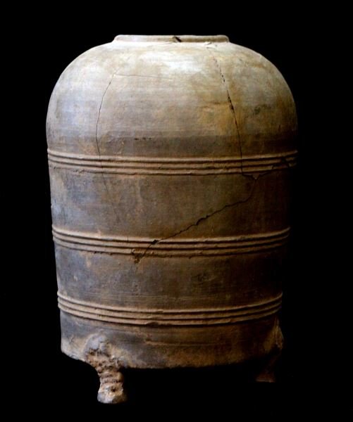 CHINESE HAN DYNASTY CYLINDRICAL CERAMIC GRANARY: Han Dynasty (206 BC to 220 AD) Decorated with thin bands of horizontal ribbing and placed upon three legs. The vessel has a hole in the bottommost band. The item has some cracks and chips but is in ov