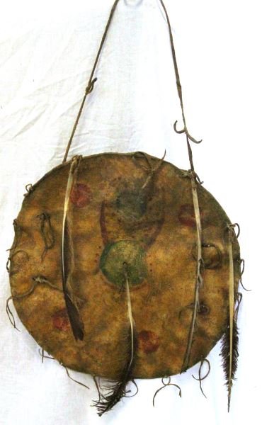 PAINTED BULL MOOSE RAWHIDE CHEYENNE SHIELD: Bull Moose rawhide with brain tanned thongs, sinew bound feathers and burned perforations, all clay paint mythical motif, superb piece age uncertain, measures 14 inches. This lot has a reserve.