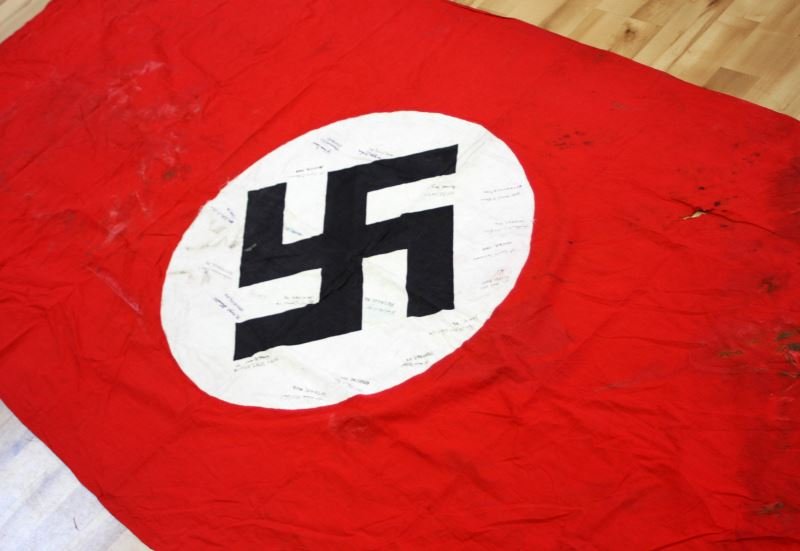 LARGE NAZI BANNER WITH CAPTURE NAMES & HOMETOWNS: 5 by 11 foot double sided Nazi swastika banner made in 3 parts. the black swastika is printed on the white discs which are then attached to the red field on either side. On one side the white disc fie