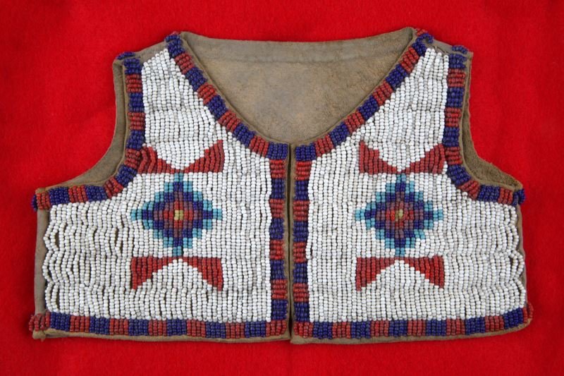 SMALL NATIVE AMERICAN BEADED VEST IN DISPLAY CASE: Small Native American beaded vest in display case, Mid-20th Century. Brain tanned leather with small beads, floral motif, only beaded border to back side. Vest measures 8.25 by 5 inches. Case measures