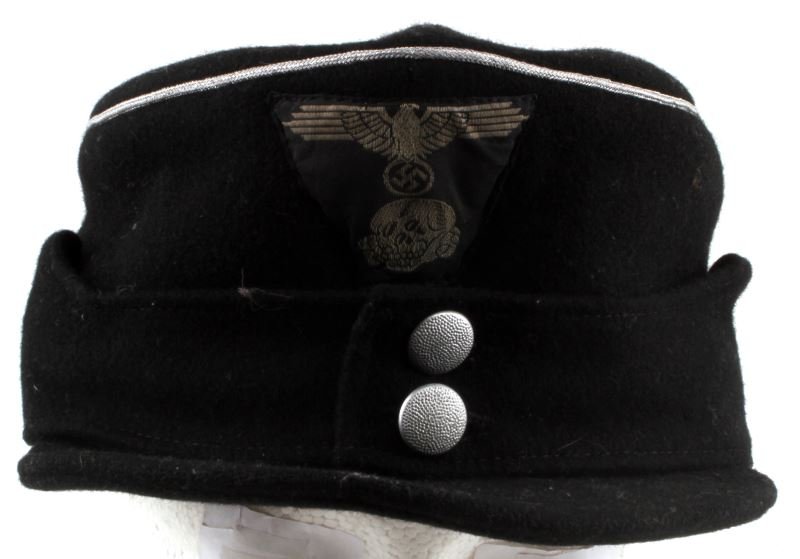 WWII THIRD REICH BLACK WOOL M-43 SS CAP: WWII German black wool M-43 SS cap. Has an SS iron eagle and a totenkopf image on the face of the cap. Silver piping. Inner leather hat rim shows degradation and is missing small chunks. A label with 