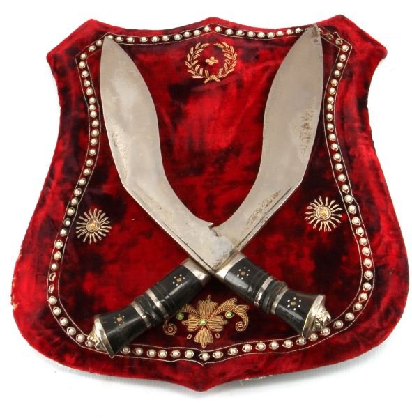 PAIR OF ANTIQUE GHURKA KNIVES MOUNTED TO SHIELD: Set of two antique Ghurka knives originating from Nepal mounted onto a red-felt cloaked decorative shield. Knives have a dark animal bone handle presumably water buffalo, knives also bear a lions head