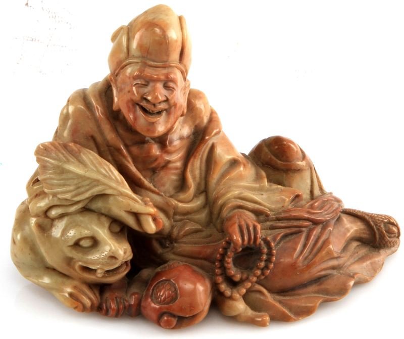 ANTIQUE CARVED SOAPSTONE FIGURINE CHINESE DEITY: Antique carved figurine, possibly depicting Zhang Guolao, made from a green and brown soapstone. The seated robber figure leans on a lion and holds beads in one hand and a feather on the other. His fa