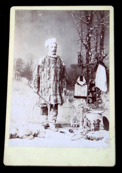 ANTIQUE CABINET PHOTO PACIFIC NORTHWEST ARTIFACTS: From a Chicago photograph firm Melander & Bros. A most unusual photo, in a seal suit, holding a superb lithic spear in one hand a mask in the other and surrounded by all manner of pacific Northwest ar