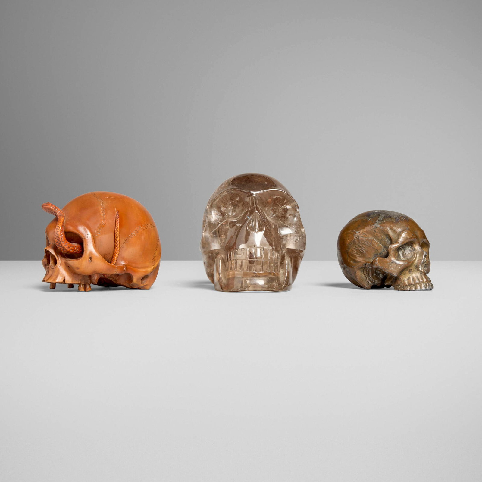 Collection of three miniature memento mori skulls: Collection of three miniature memento mori skulls carved wood, smoky quartz, bronze 3.25 h x 2.75 w x 4.25 d in (8 x 7 x 11 cm) 2.75 h x 2.75 w x 3.5 d in (7 x 7 x 9 cm) 2 h x 2.25 w x 3 d in (5 x 6
