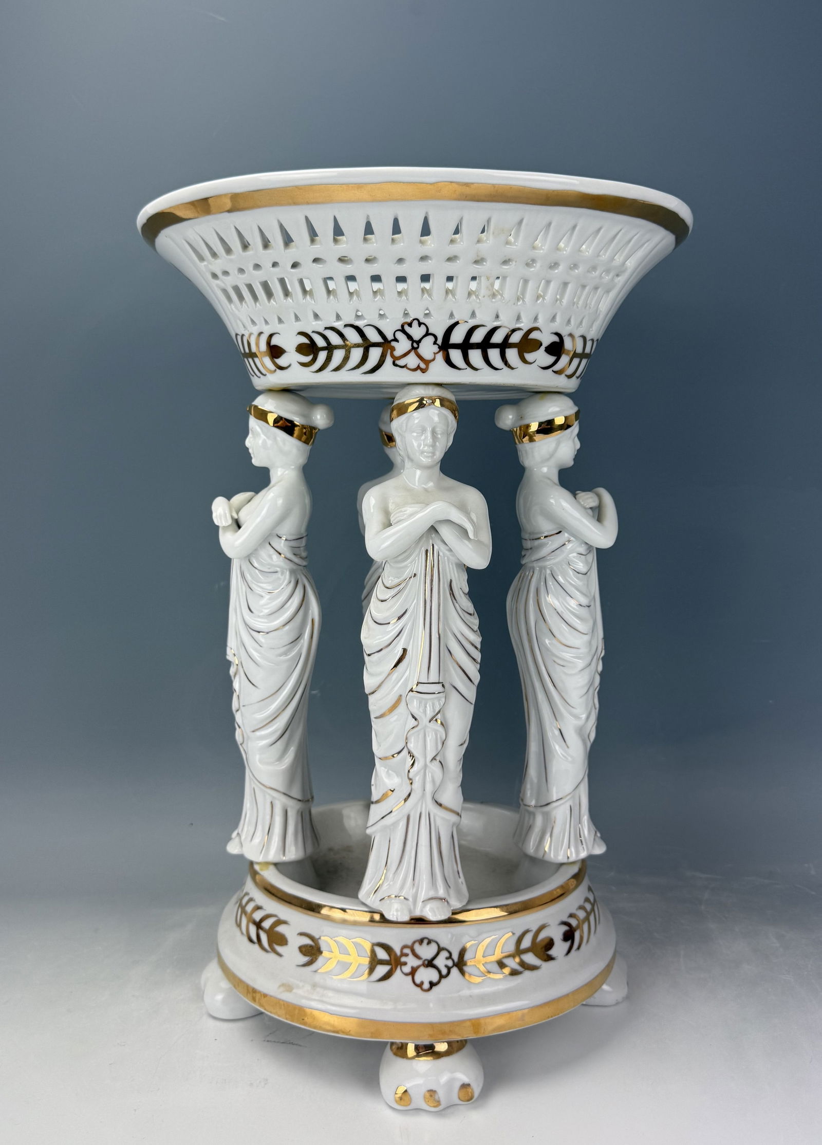 A large Vintage Porcelain Tazza: A large Vintage Porcelain Tazza,Three graces in bisque porcelain decoration standing on a postament carrying a pierced basket on their head. H 40 cm (17.5in.) x W 29 cm (12in.) x D 26 cm (10 in.). Pre