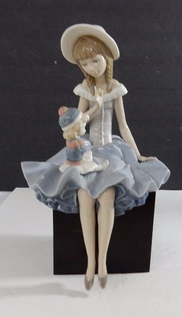 Lladro Porcelain "Talking To The Doll" Figurine: DESCRIPTION: A Lladro porcelain figurine titled: "Talking To The Doll", item number: 01001378. Features a young, elegantly dressed girl, seated on a wooden cube, talking to a doll perched atop her lap