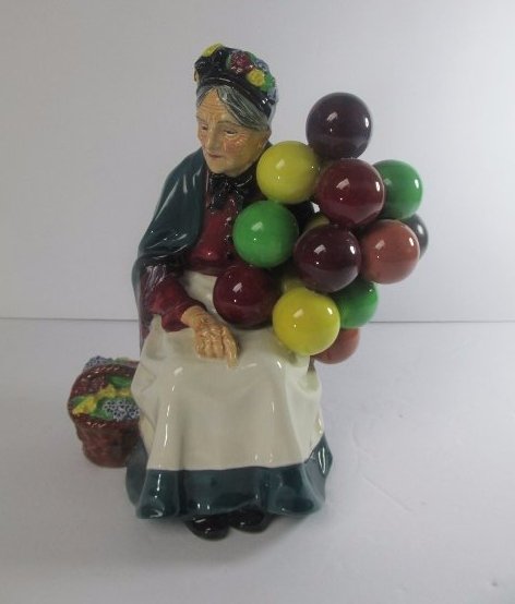 Royal Doulton "The Old Balloon Seller" Porcelain: DESCRIPTION: A Royal Doulton porcelain figurine titled: "The Old Balloon Seller". Features an elder woman seated in a bench with a basket by her side, as she grasps a group of floating balloons. Marke