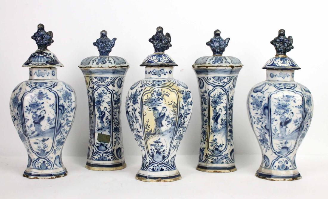 Dutch Delft Blue and White Five Piece Garniture: Three of faceted baluster form with raised lid and figural finial, two of beaker form decorated with lobed panels depicting a Chinese figure bearing a flower filled vase within foliate ground. Extensi