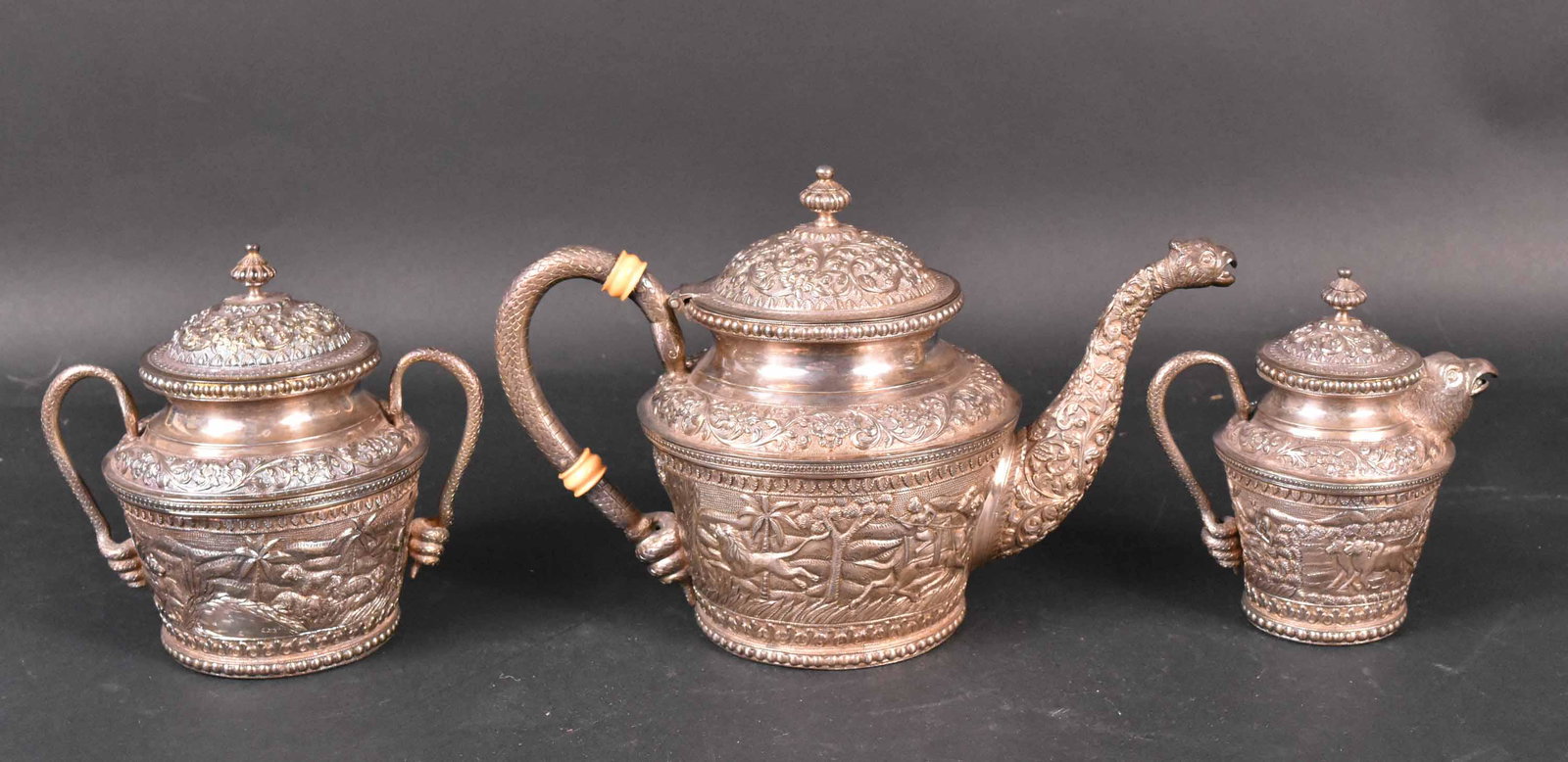 Pakistani Silver Three Piece Tea Service: Makers Mark J. Manikrai Karachi T.95, monogrammed on underside RVP, densely chased scroll floral, hunting scene with palm trees, snake handles, animal figural spout, comprising teapot, covered sugar b