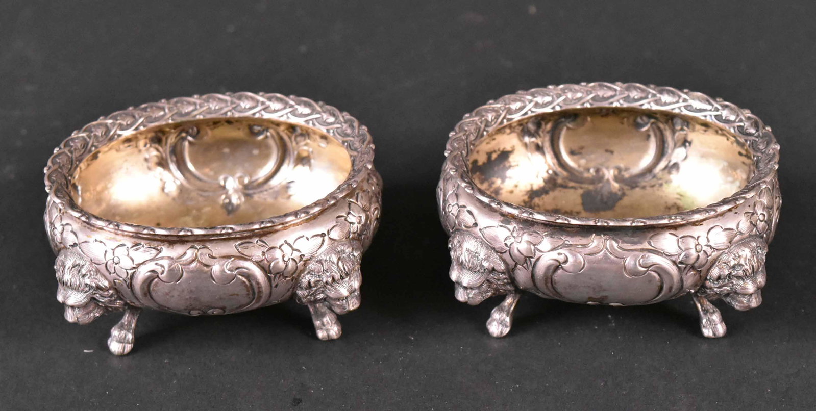Pair of Wood & Hughes Silver Salt Cellars: Makers Mark, W&H, Wood & Hughes, New York, Circa 1875, oval heavily chased with intricate looped border, floral & scroll motif, each side with vacant cartouche, each raised on lion mask and foot, each
