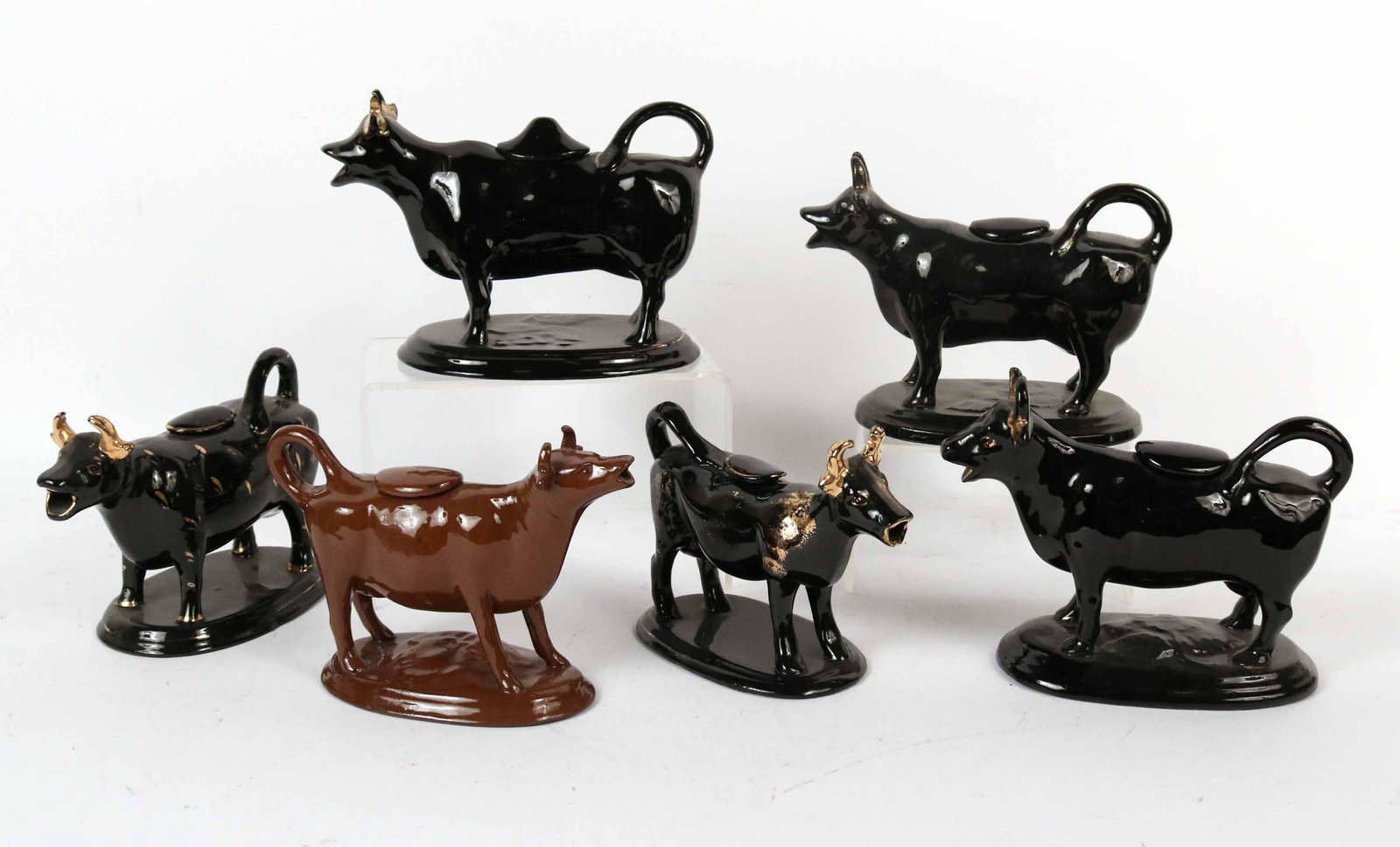 Five English Jackfield Earthenware Cow Creamers: Decorated with a black glaze and gilt decoration, all five on an oval plinth; the upturned tails form the handle, with fitted lids on the backs. Together with an English Staffordshire brown glazed cow