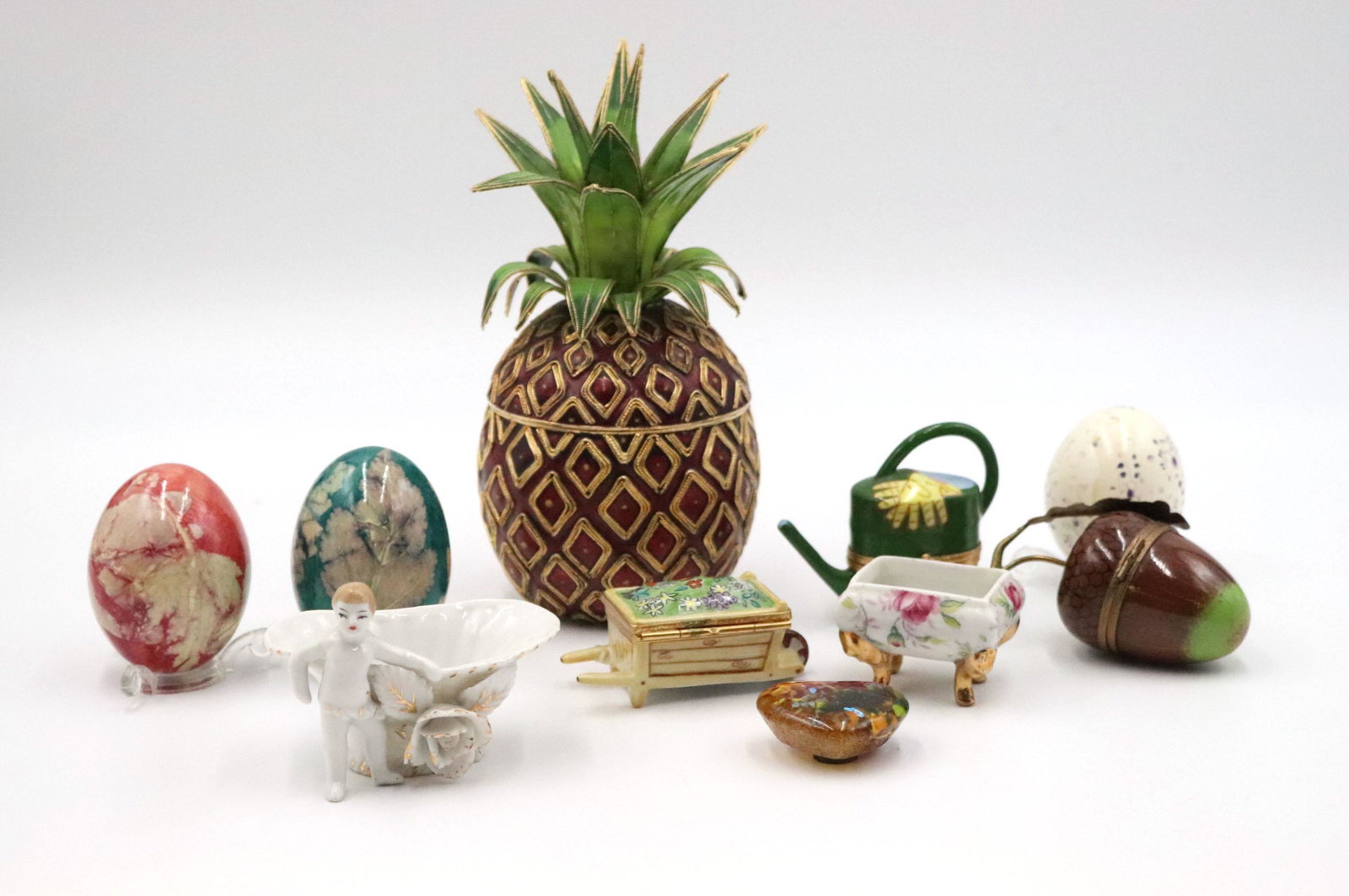 Two Limoges Figural Porcelain Pill Boxes: Together with a art glass magnet, three eggs on stands, an enamel pineapple form box, a acorn form pill box, a basket with putti, and a floral decorated box on feet. 10 pieces Cracks and wear. Pineapp
