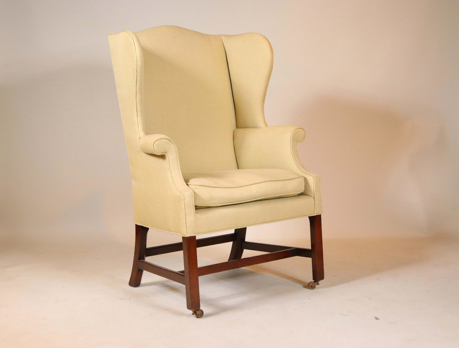 George III Style Upholstered Wing Armchair: The padded serpentine backrest with shaped wings above a loose-cushioned seat flanked by outscrolled armrests, raised on chamfered square legs joined by stretchers, the front feet on casters 43-1/2"h 