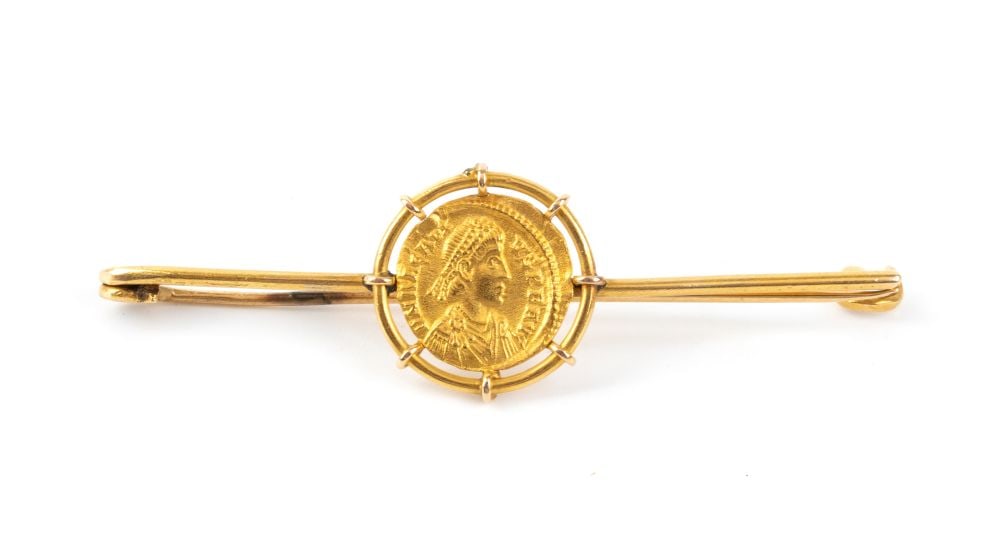 Gold brooch with a coin: Elegant yellow gold brooch executed in the Roman Archaeological Revival style, featuring a slender linear mount supporting, by means of six evenly spaced claws, a reproduction of a Late Roman Imperial