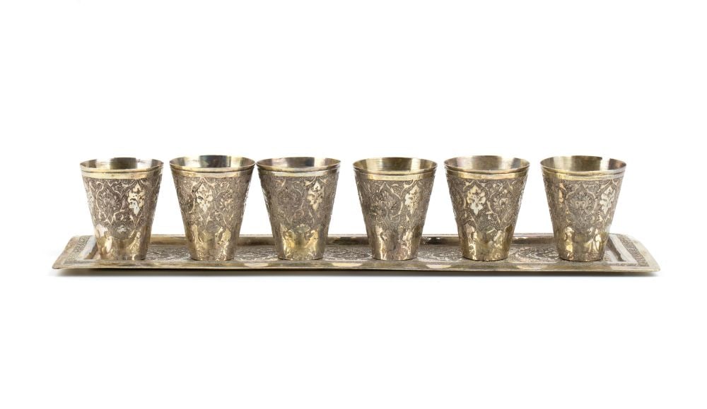 A group of 6 solid silver glasses and a silver tray: comprising six silver glasses with surfaces engraved with plant and floral motifs, resting on a rectangular tray richly engraved with the same floral and plant motifs.Height x width of saucer: 27.7 x