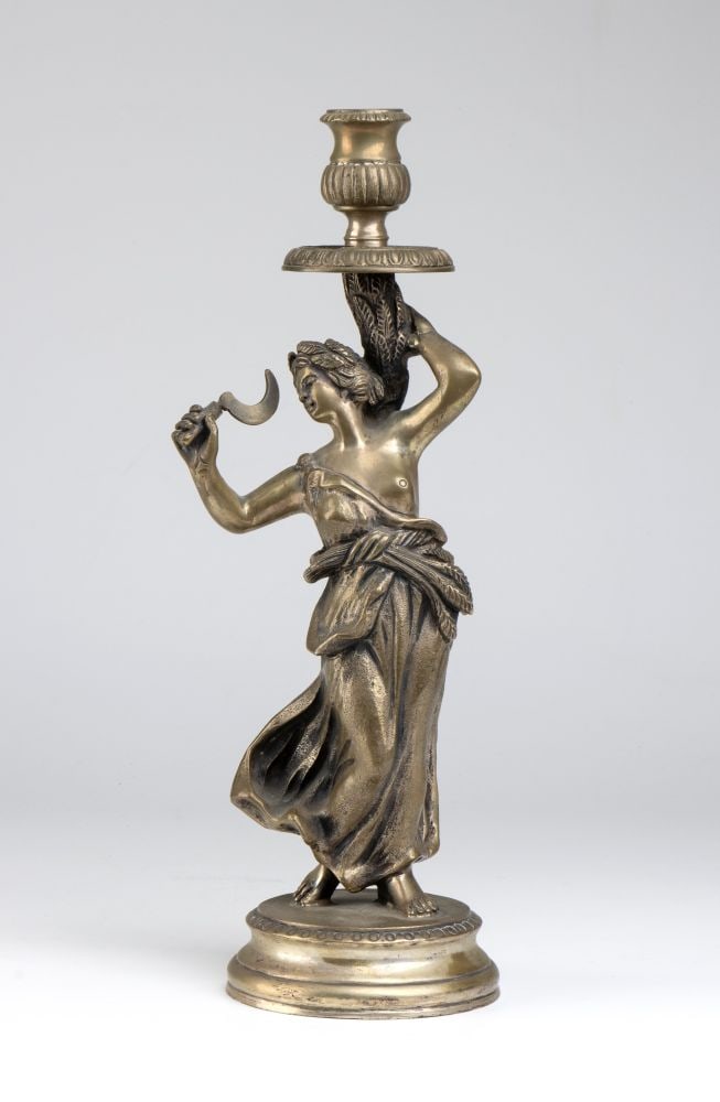 Italian solid silver candlestick: cast in fusion. It rests on a circular base from which rises a female figure in classical dress; in her right hand she holds a sickle, whilst with her left she raises a sheaf of wheat, symbolising the