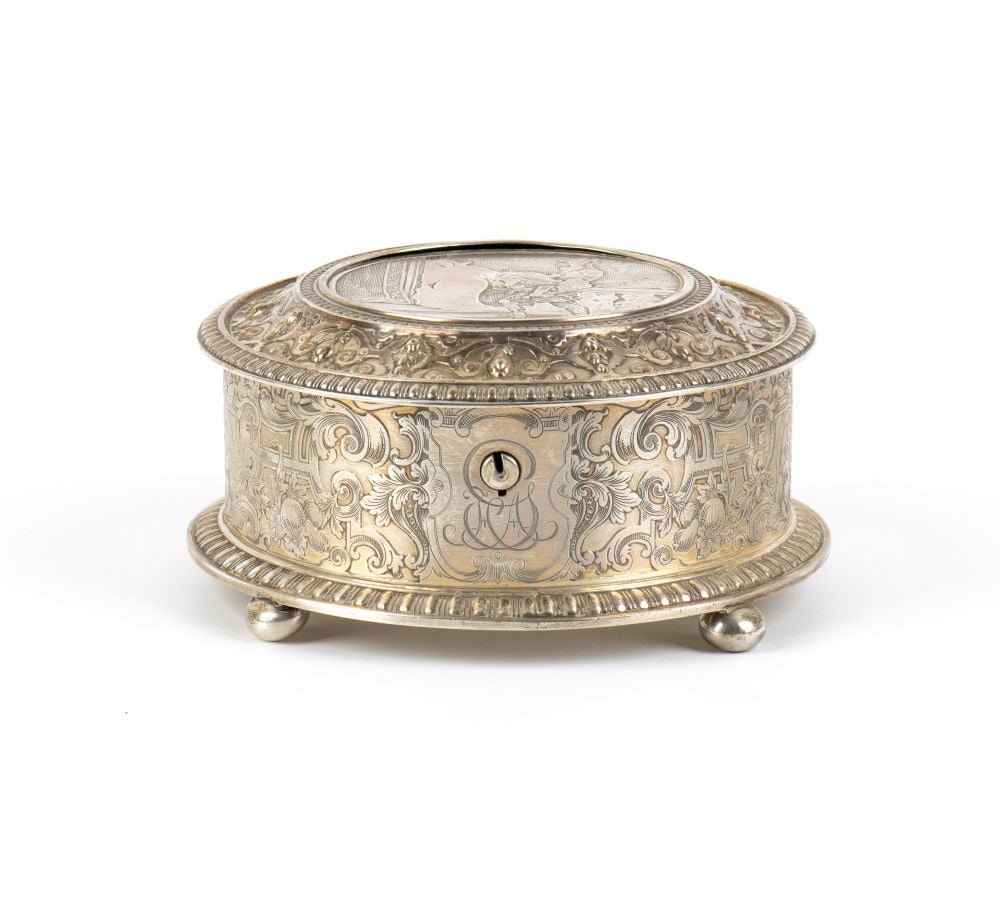 REED & BARTON American sterling silver jewellery box: circular in shape, resting on four spherical feet. The body is decorated with dense motifs of acanthus scrolls and architectural elements. The lid is hinged and lockable, flanked by a monogram with