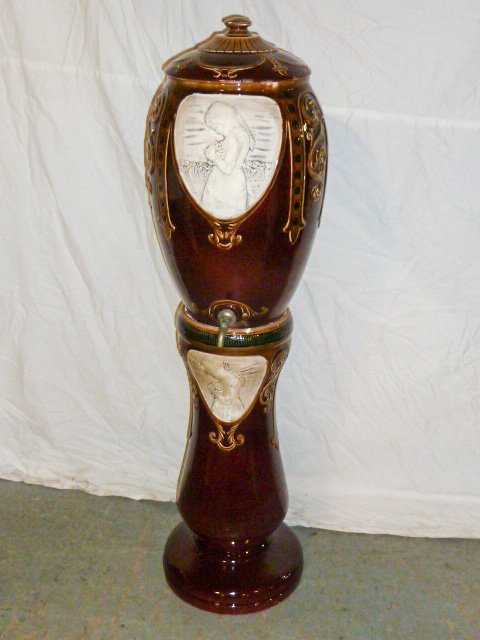 Arts & Crafts Era Majolica Water Cooler & Pedestal: High glaze finish , Center medallions " Girl gathering flowers" . Original lid . Measures 42 1/2" tall. Cooler measures 19" and Pedestal 23 1/2" . As found estate condition . Please use photos as part