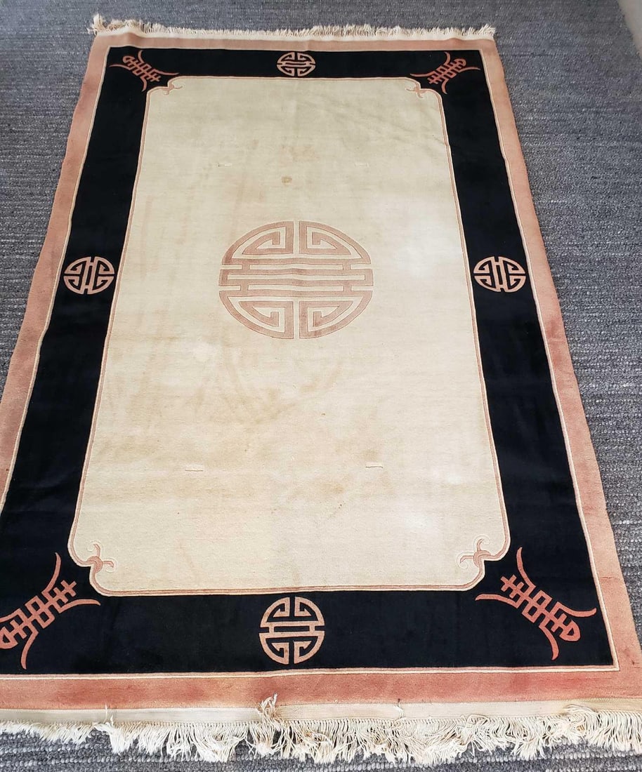 VINTAGE CHINESE ART DECO SHOU WOOL RUG IN IVORY BLACK AND TAN: This vintage handmade wool Chinese Art Deco rug, measuring 5 feet 11 inches by 8 feet 8 inches, showcases a classic design with a deep beige field and a striking blue border. The rug's central motif f
