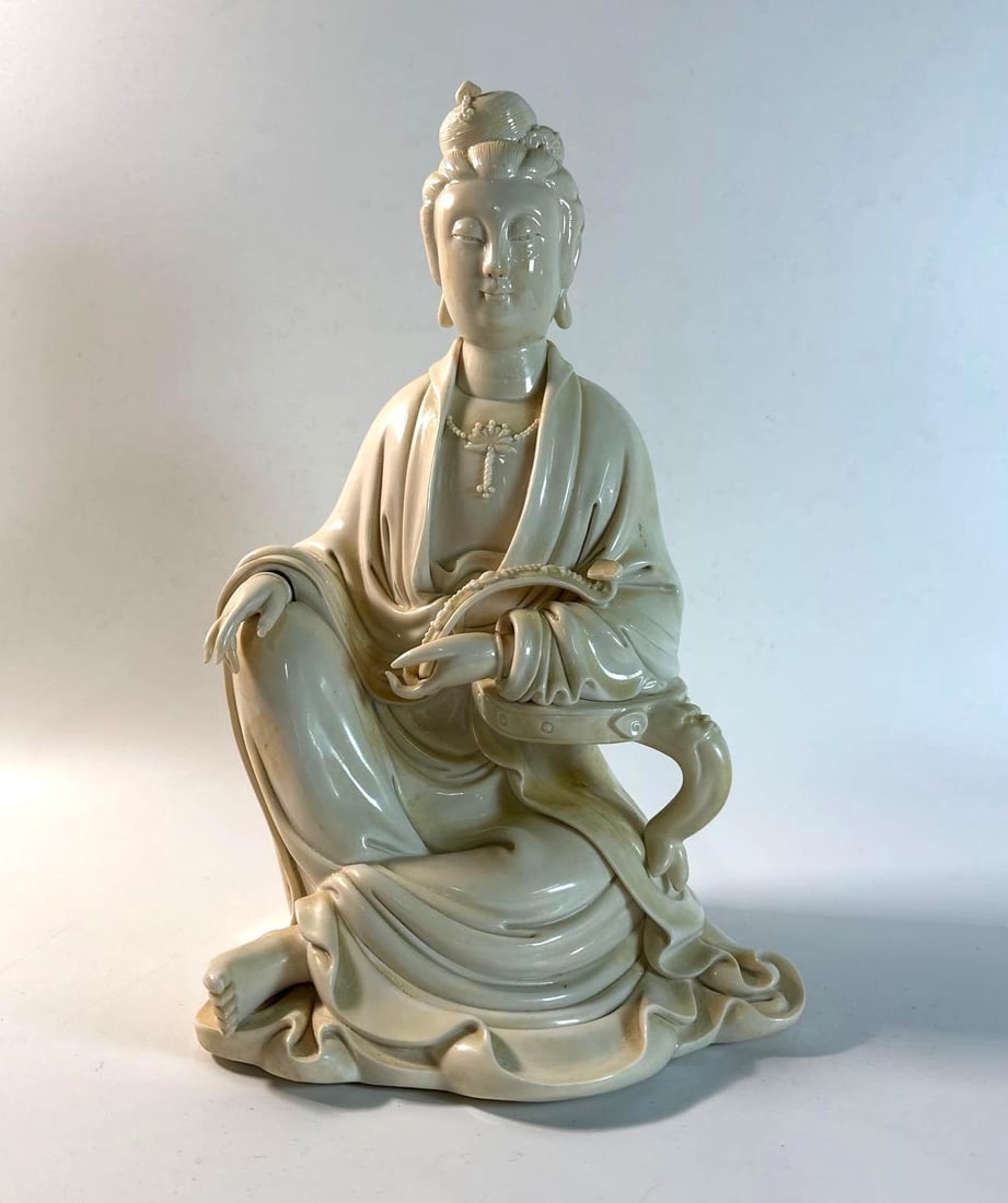 CHINESE BLANC DE CHINE DEHUA PORCELAIN SEATED GUANYIN STATUE: This Chinese Blanc de Chine Dehua porcelain statue, standing 14 inches tall, depicts a seated Guanyin, the bodhisattva of compassion, showcasing the serene beauty of Buddhist art and the refined craft