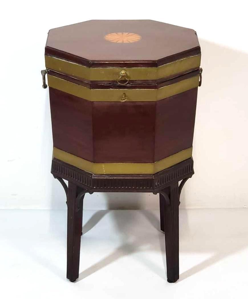 GEORGE III MAHOGANY BRASS BOUND INLAID CELLARETTE: A George III mahogany brass-bound inlaid cellarette, featuring an octagonal shape with brass banding, inlay details, and a hinged lid, raised on four legs. Dimensions: 29"H X 17.5"W X 17.5