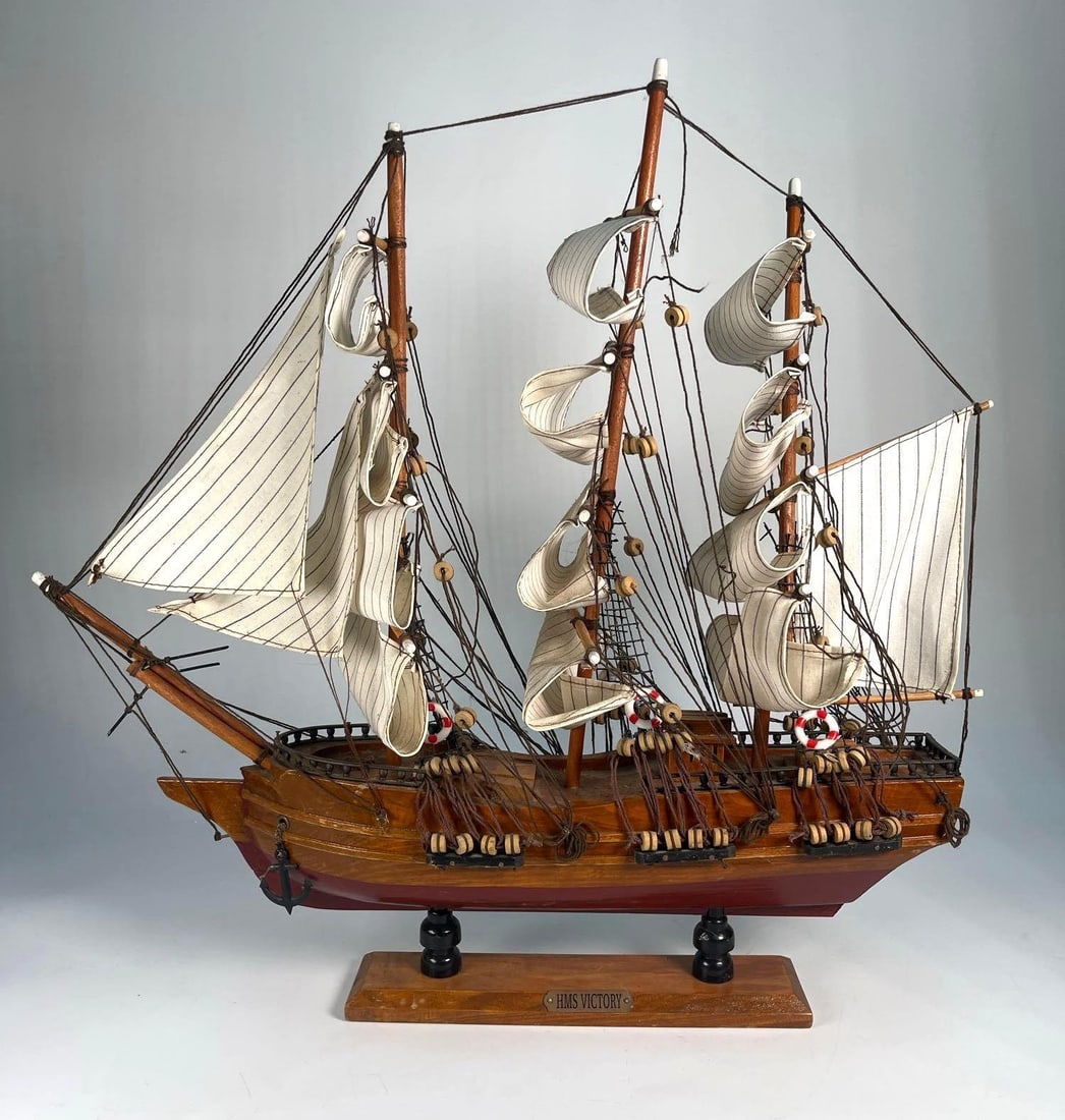 WOODEN HMS VICTORY BOAT: A detailed wooden model of the HMS Victory, a famous British Royal Navy ship, is displayed with its masts, rigging, and partially furled sails accurately represented, mounted on a wooden base with a p