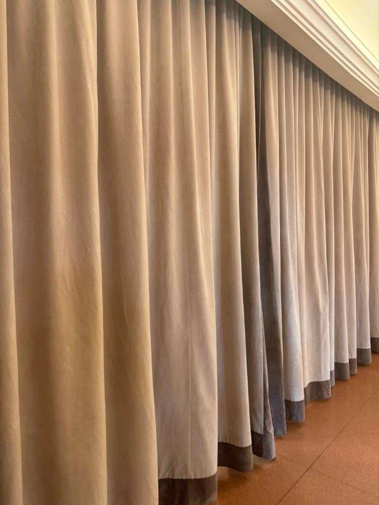 PAIR OF TAUPE VELVET BLACKOUT CURTAINS WITH BROWN TRIM: A pair of beige blackout curtains, each featuring a brown trim along the bottom. From the Moschino NYC showroom. Dimensions: 10' x 165", 10' x 72" (2 panels) Artist or Maker: Custom Made This item