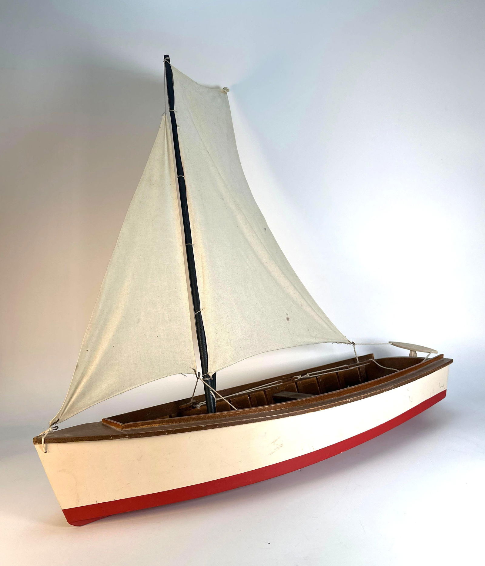 WOODEN SAILBOAT MODEL: A handcrafted wooden model of a sailboat is shown, featuring a single mast with sails, a rudder, and a painted hull with red accents. Dimensions: 24” Tall x 24” L x 9” W Artist or