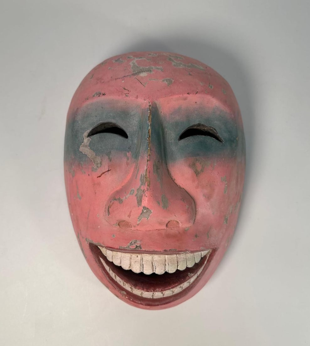 RARE INUIT MASK: A carved, wooden mask with a pink base, blue shading around the eyes, and a wide, open-mouthed grin revealing a full set of teeth. Dimensions: 10"L X 8.5"W Artist or Maker: Unknown Provenance: