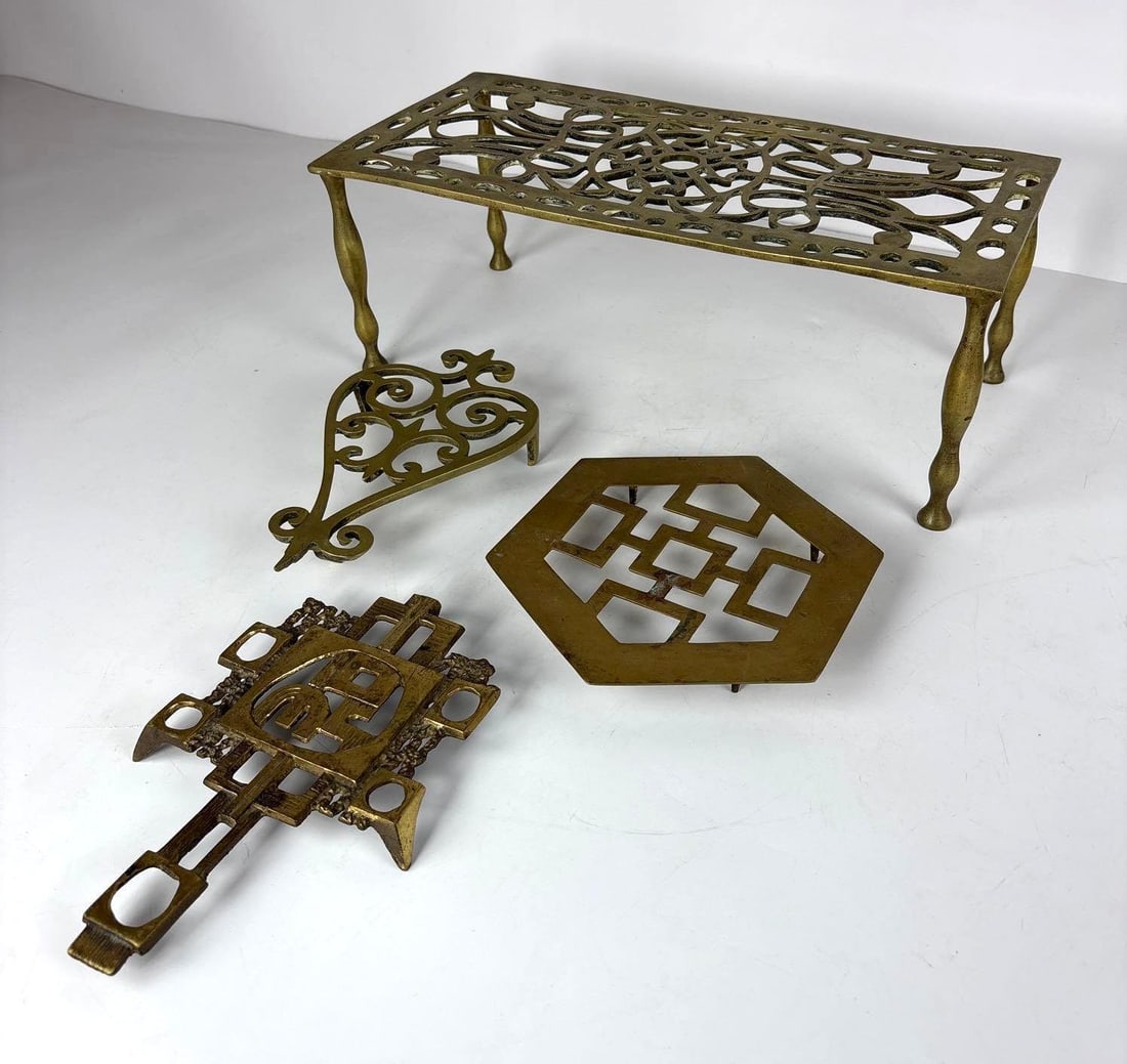 BRASS TRIVET LOT: A group of four decorative brass items, including a long rectangular plant stand or trivet, a triangular scroll-work trivet, a hexagonal trivet with an intricate geometric pattern, and a brass cross-s