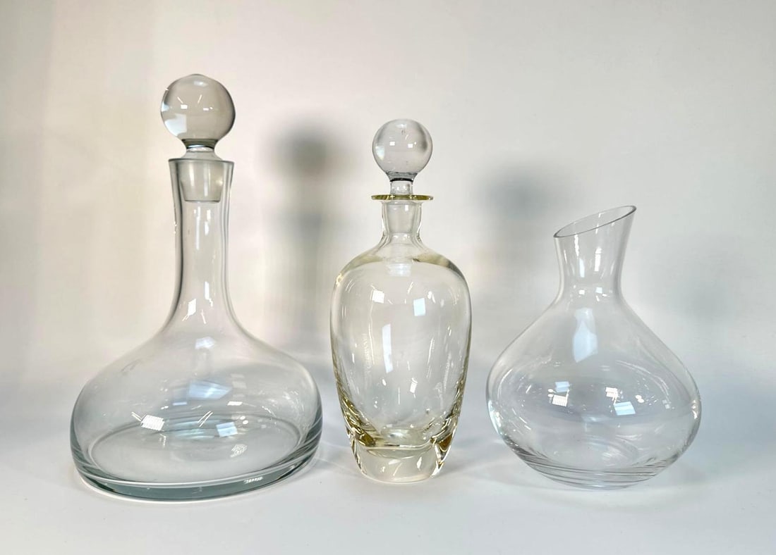 LOT OF VINTAGE DECANTERS: Three vintage clear glass decanters, with two having spherical stoppers and one being a squat, open-top carafe with an angled rim. Largest Dimensions: 12"T x 7.75"DIA Artist or Maker: Unknown 