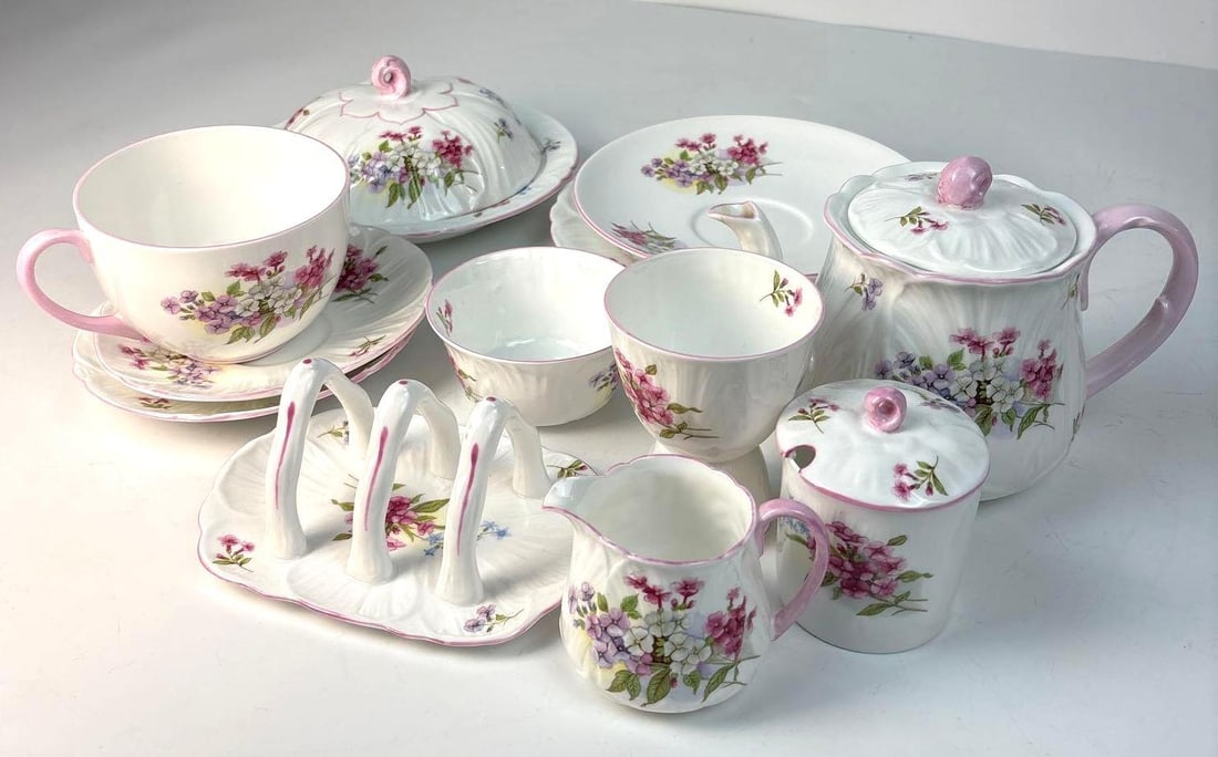 SHELLEY STOCKS TEA SET: *A collection of white porcelain tea and serving ware, featuring pink-rimmed edges and delicate floral illustrations in shades of pink, purple, and green. Dimensions of Largest: 8" Diameter Artist